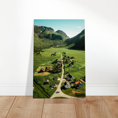 Aerial view of a quaint countryside village surrounded by rolling green hills and distant mountains under a clear sky.