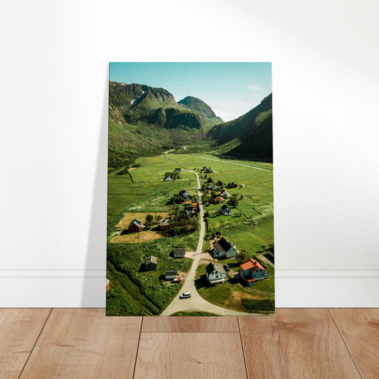 Aerial view of a quaint countryside village surrounded by rolling green hills and distant mountains under a clear sky.